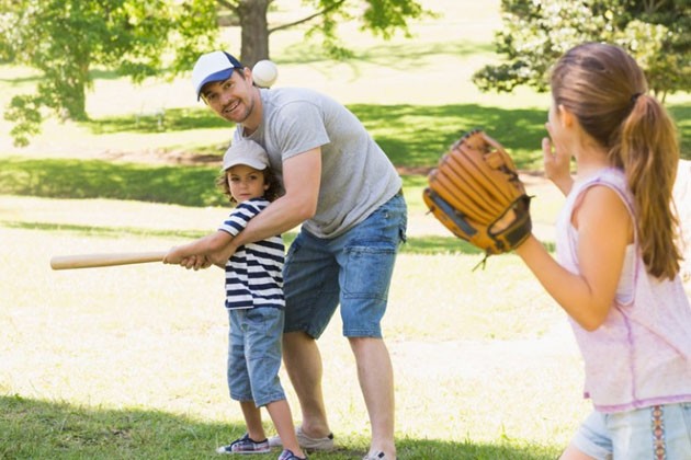 An older child throws a baseball while the father helps a younger child swing the bat.