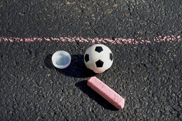 A small plastic bottle cap, stick of chalk and small ball sit below a chalk line drawn on the pavement.