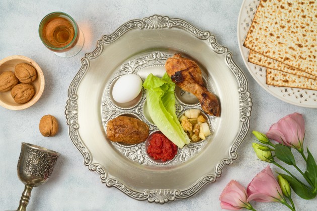 A silver plate full of traditional Passover foods, with matzo, walnuts and flowers nearby on the table