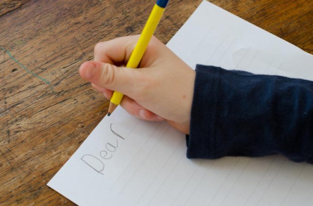 A child’s hand holding a pencil poised over a sheet of paper with the word “dear” on it.