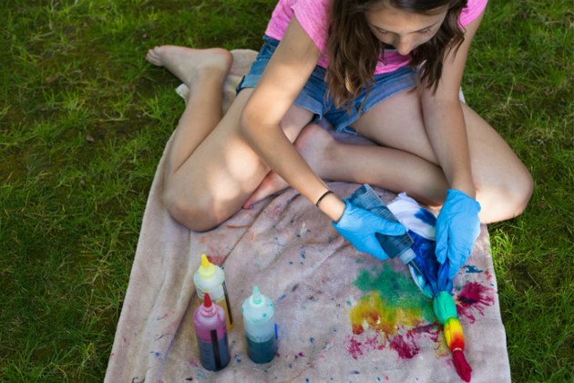 A child wearing gloves sitting on a tarp and squeezing dye to create a tie-dye shirt.