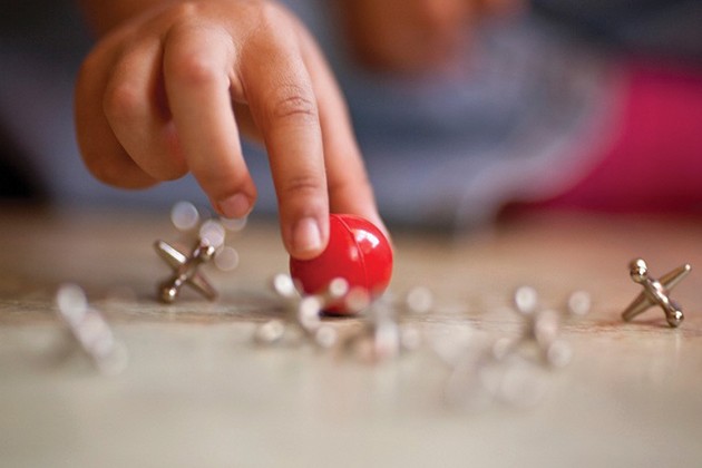 Child’s fingers holding a rubber ball near jacks on a table.