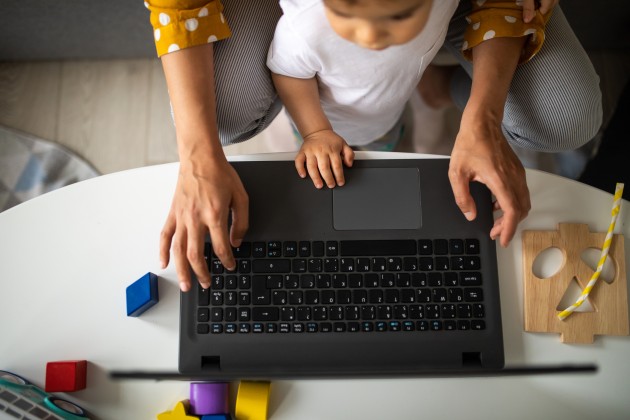 Woman with baby on lap using laptop on a desk with kids’ toys.