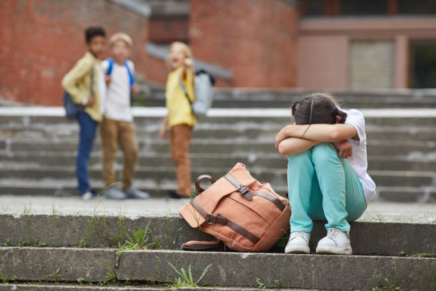 Little girl sitting on steps with her head in her lap while a group of kids points and laughs.