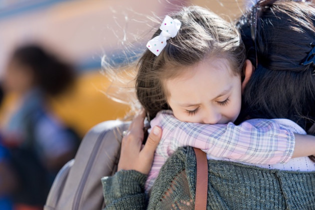 Little girl hugging mother before school with school bus in the background.