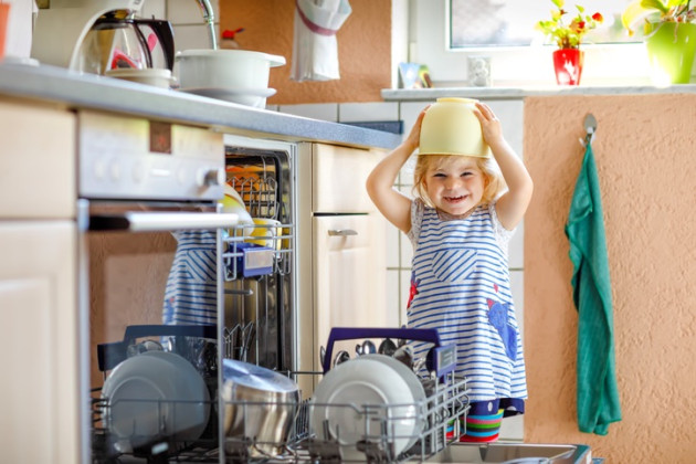 Funny happy toddler girl standing in the kitchen, holding dishes and putting a bowl on head.