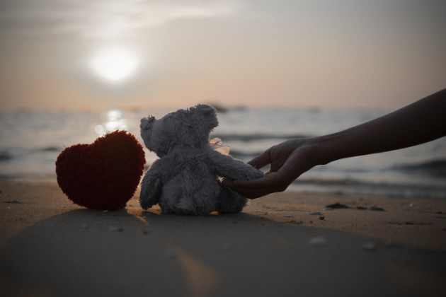 Stuffed teddy bear and crocheted heart on beach with woman’s hand.
