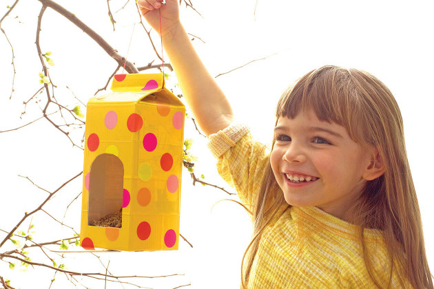 A grinning child proudly holding up a birdfeeder made from a paper carton.