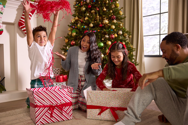 A family of four sits in front of a decorated Christmas tree, opening presents and smiling, with one child excitedly throwing gift wrapping in the air, enjoying a stress free holiday together.