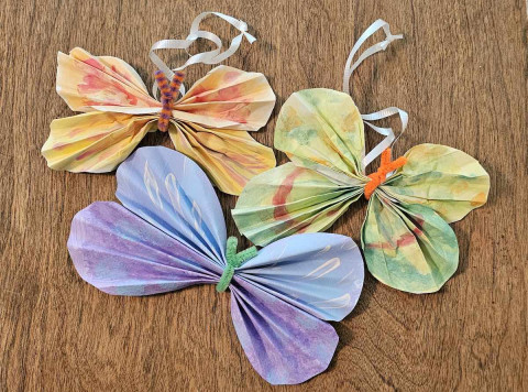 Three vibrant paper butterflies with ribbons resting on a wooden table surface.